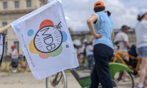 Ee arrière plan, une cycliste est prise de dos à côté de son vélo, au premier plan, un drapeau aux couleurs de MDB