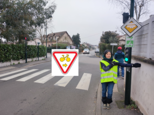 Au pied d'un feu rouge, une cycliste avec un gilet jaune indique avec son bras tendu la droite. Sur sa gauche un panneau M12 Droite